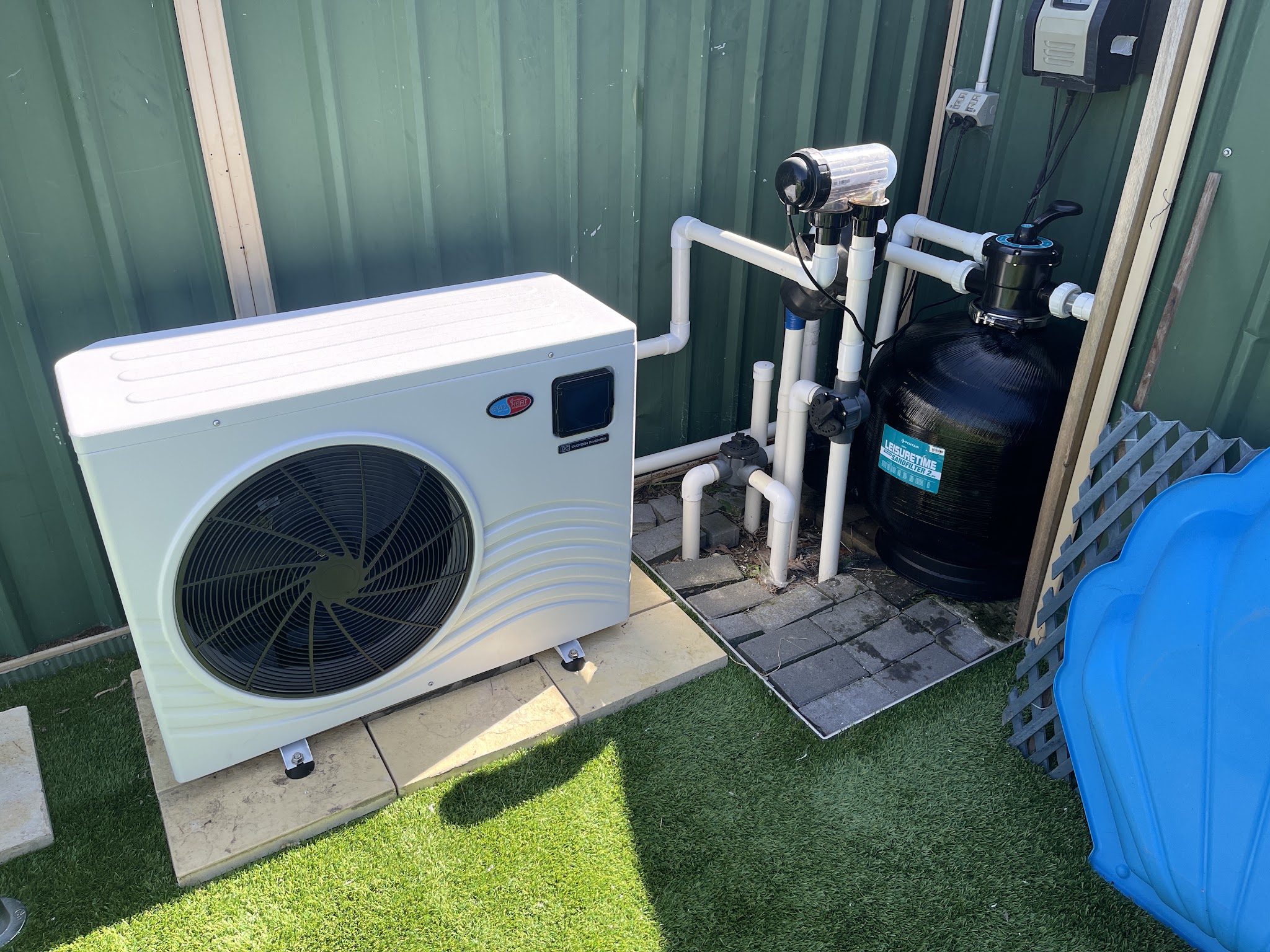 A white heat pump and sand filter installed side by side against a green fence